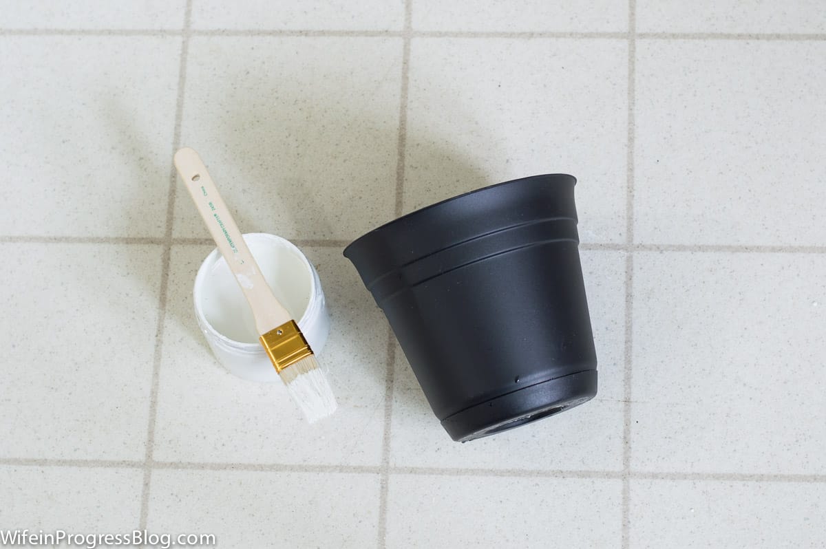A black, plastic bucket with a can of white paint and paintbrush nearby