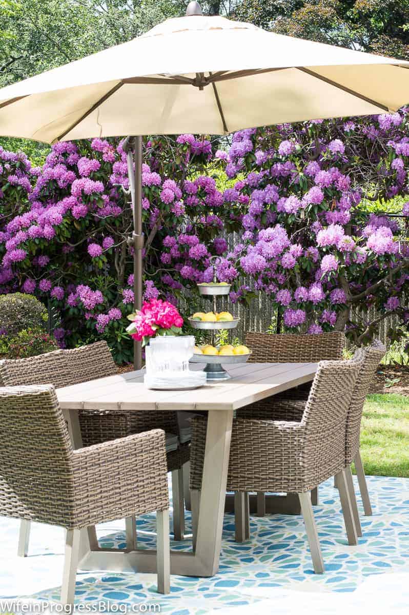 A beige umbrella on a light brown patio table with six wicker chairs, on blue mosaic tiles, and purple flowering shrubs in the background