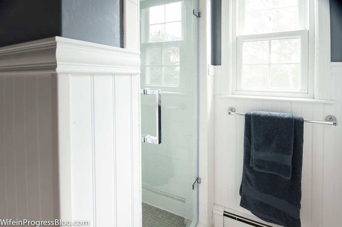 A stand-up shower with a glass door and chrome handle, near a window with a towel rack and a navy blue bath towel set