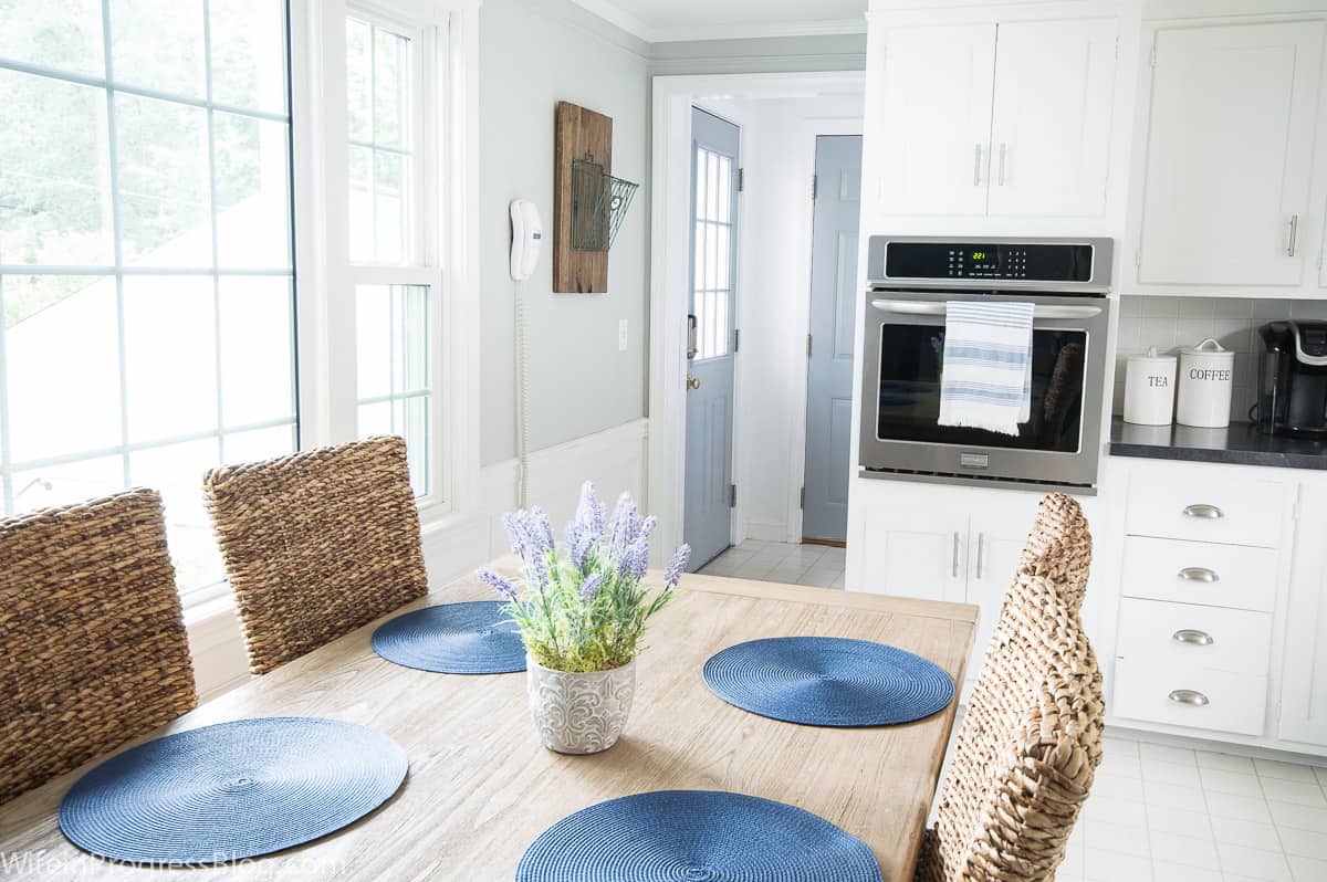 A kitchen with dining table, wicker chairs, large windows on the left, and a door leading outside (painted light blue), along with a closet door (also painted light blue)