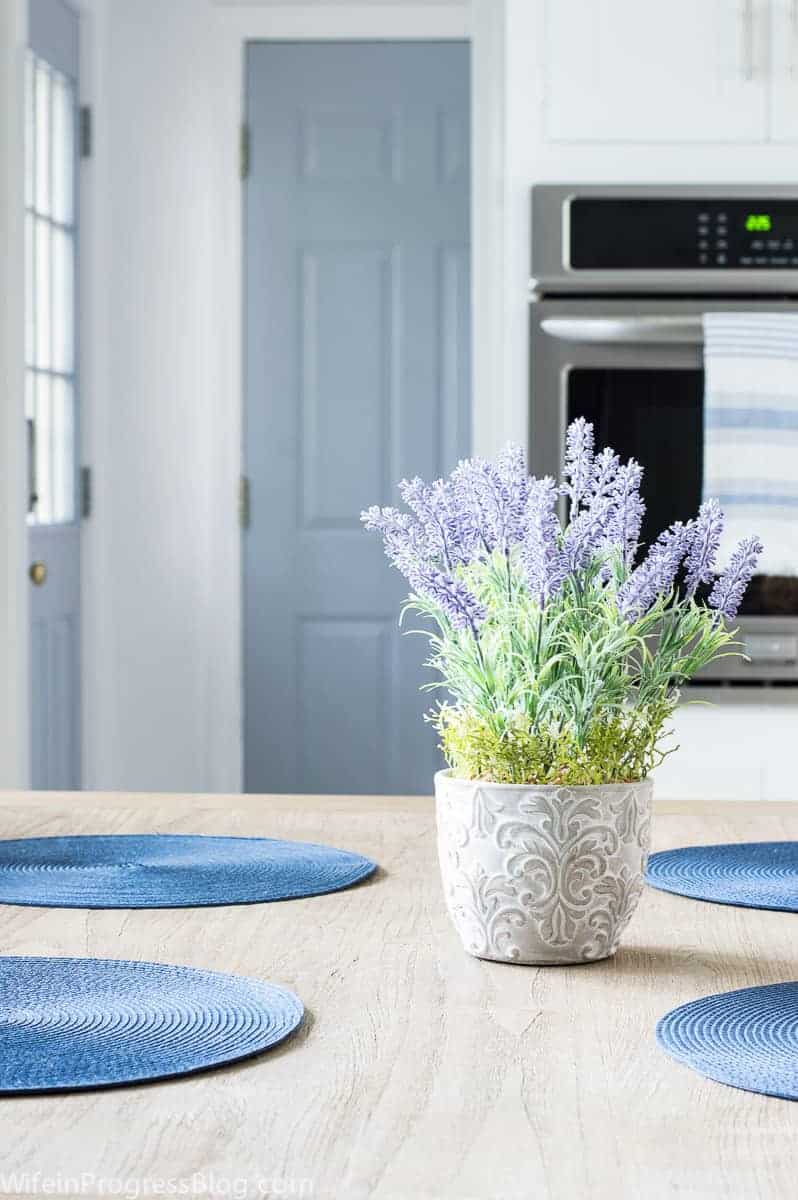 A grey and white plant pot with light purple flowers, on a wooden dining table with round, blue place mats, and a light blue door in the background