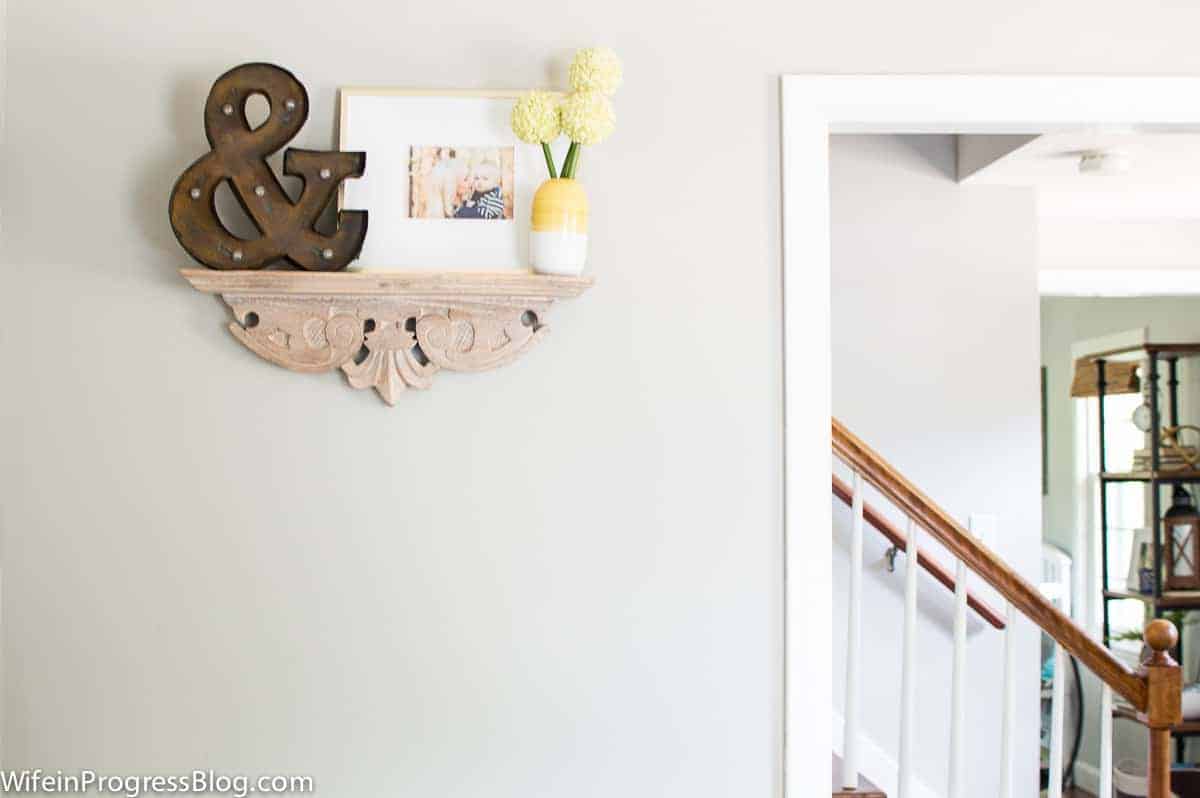 A light brown floating bookshelf with a fleur de lis design, holding a vase of yellow flowers, photo frame and wooden \"ampersand\" decor  