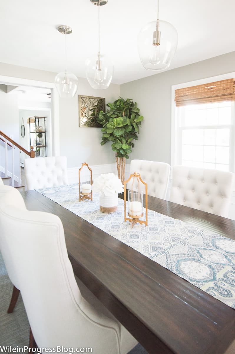 A dining room with modern, dark wooden table, blue and white table runner, grey upholstered chairs and a tall house plant in the corner