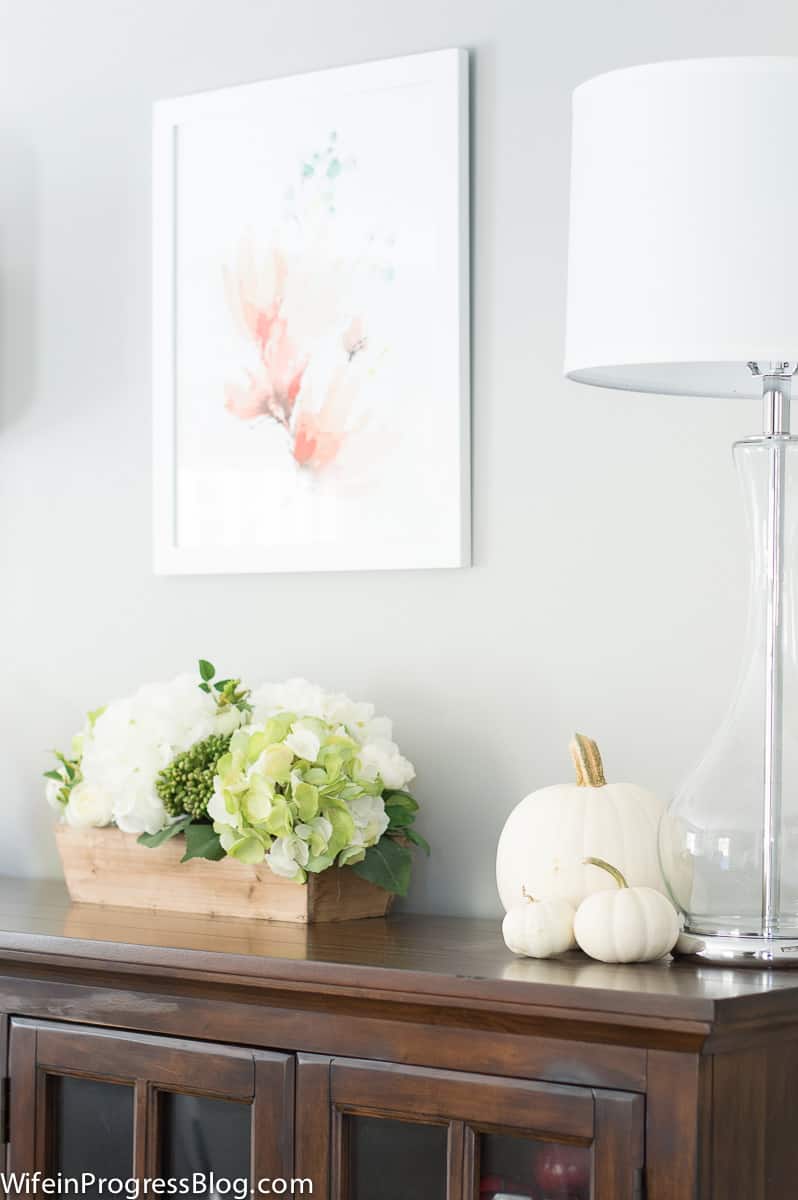 A tabletop with tall, chrome and white lamps with clear glass bases, next to off-white pumpkins of various sizes and a wooden box with green and white flowers