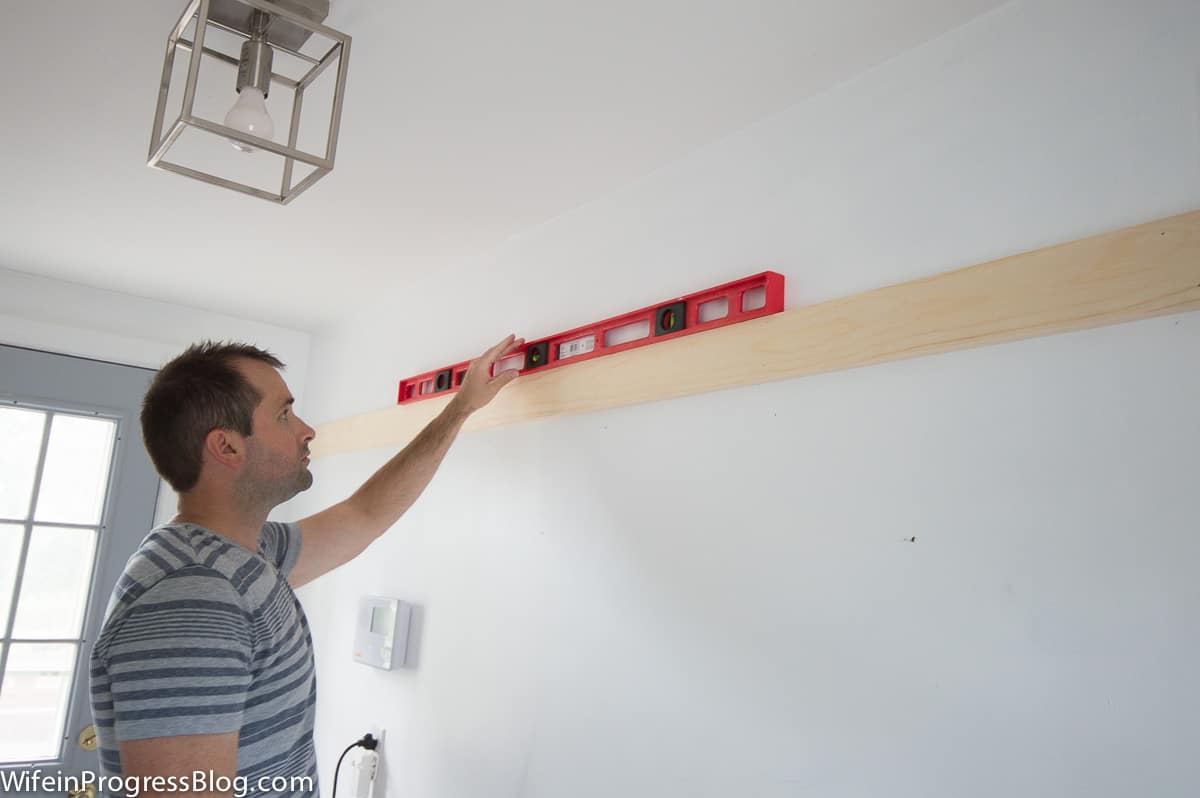 A man holding a level above a plank of unpainted wood that is now secured to the wall