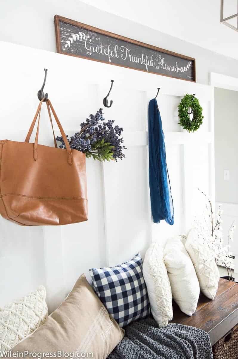 A wall featuring board and batten design, with pegs for a large, brown bag holding greenery, and a wooden bench below, with assorted pillows resting on top