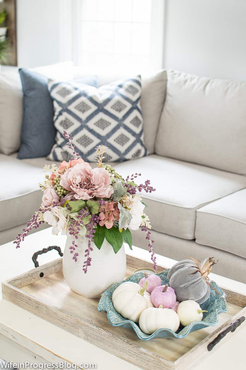 A vase of flowers, a bowl of pastel pumpkins, on a wooden tray on a coffee table, with the corner of a sofa in the background