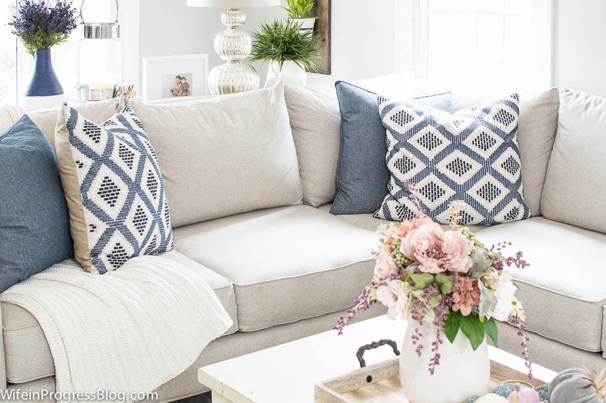 A light grey sofa with blue and white pillows, an off-white wooden coffee table holding a white vase of flowers on top