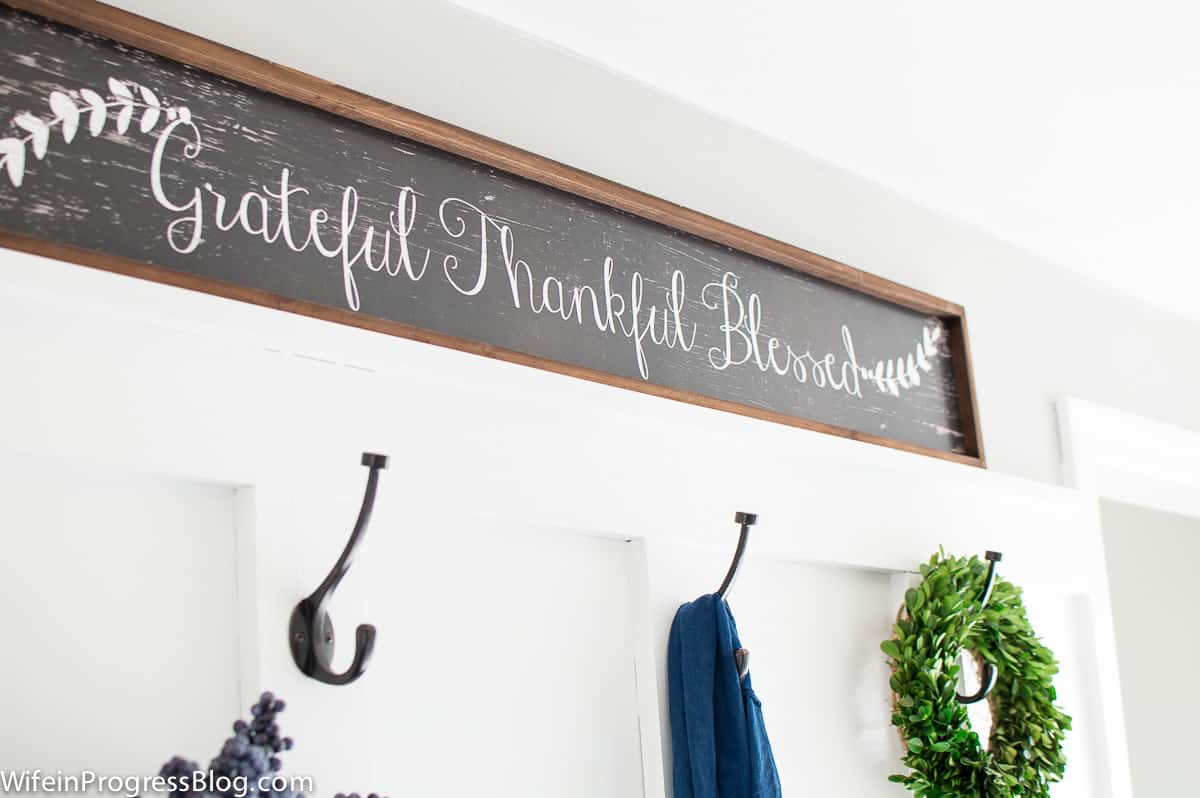 A long, rectangular chalkboard sign with the words \"Grateful, Thankful, Blessed\" resting above a row of metal pegs on the wall