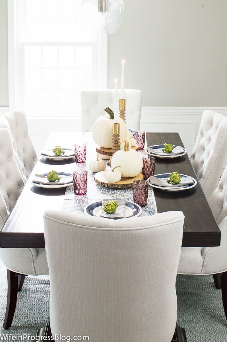 A dining room table with off-white pumpkins, blue and white place settings, green artichoke embellishments and purple glassware