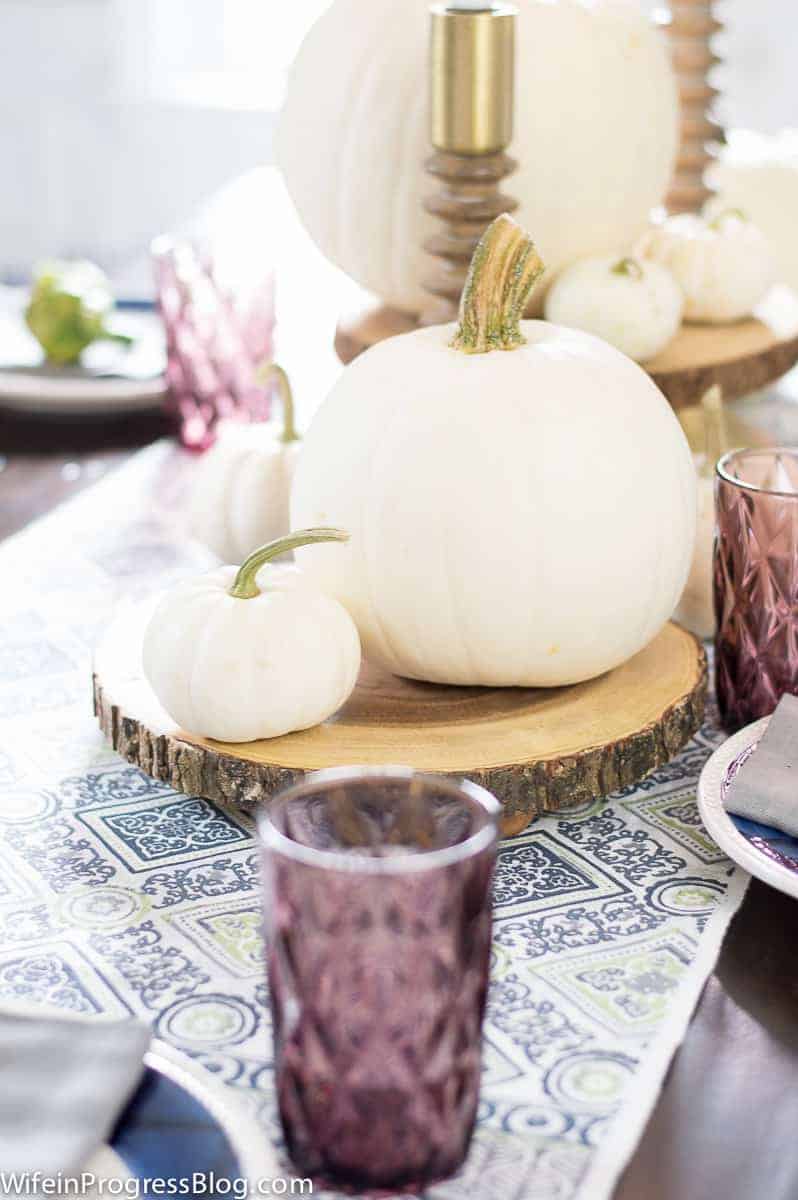 Large, off-white pumpkin with smaller ones nearby, sitting on rustic wooden circle, on a blue/white table runner and purple glasses