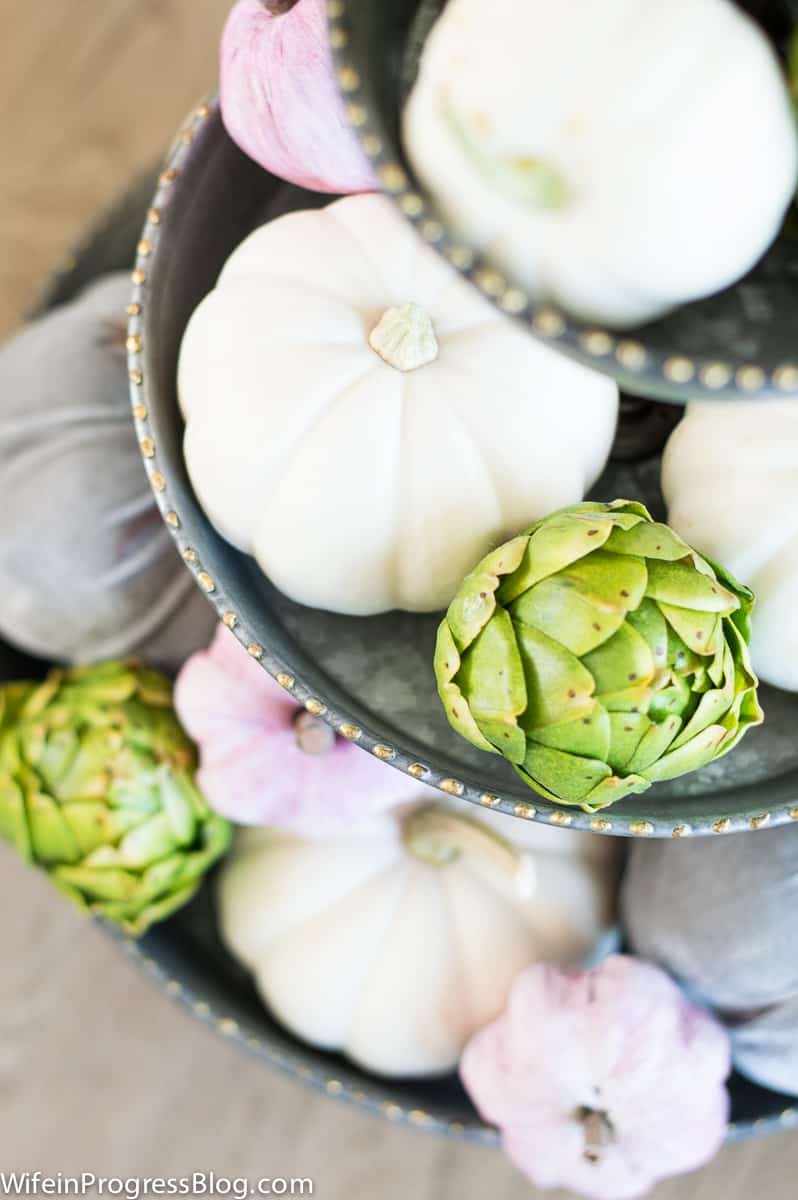 A top view of off-white, mauve and grey pumpkins and green artichokes on a metal 3-tiered tray