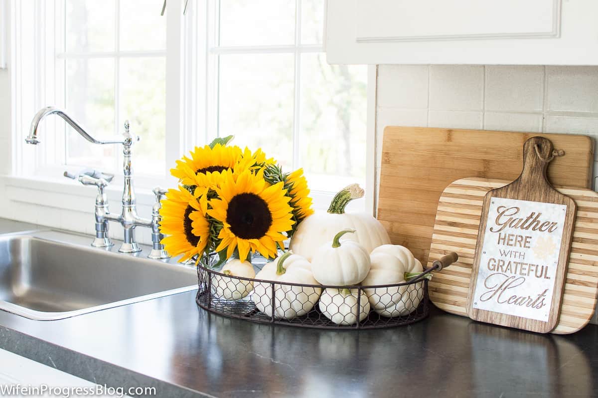 A black wire basket holding off-white pumpkins and a vase of sunflowers on a kitchen counter top near the sink and window, with cutting boards nearby