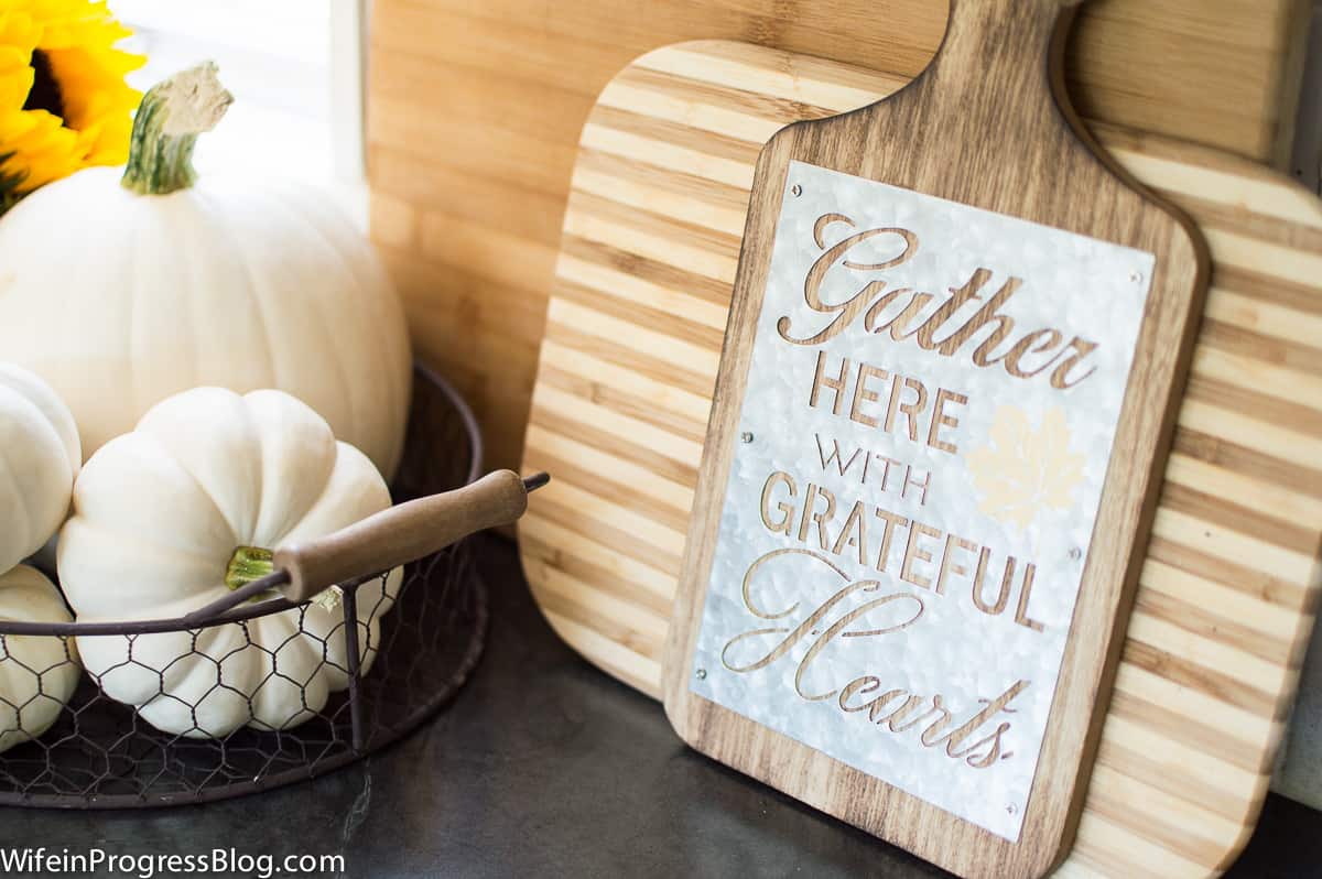 A wooden cutting board with the words \"Gather here with grateful hearts\", on a counter top with other boards and a basket of pumpkins