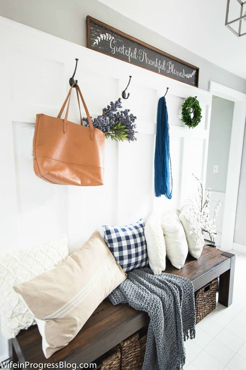 An entryway with board and batten wood detailing on the left wall, a dark bench below with wicker baskets, and various pillows on top