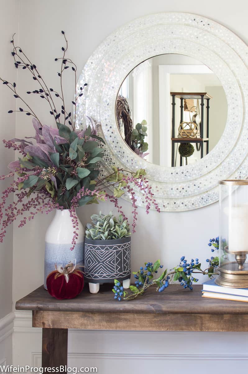 A wooden console table with a large arrangement in a vase, a potted plant nearby, miscellaneous decor and the large, round mirror above