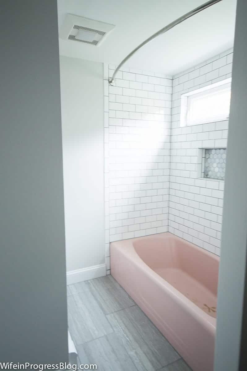 A pink tub, white subway tile around tub walls, an oblong window above and a curtain rod