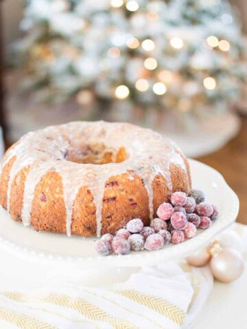 A bundt cake with glaze on top, and frosted cranberries on the side, on a white cake stand, with a lit Christmas tree in the background