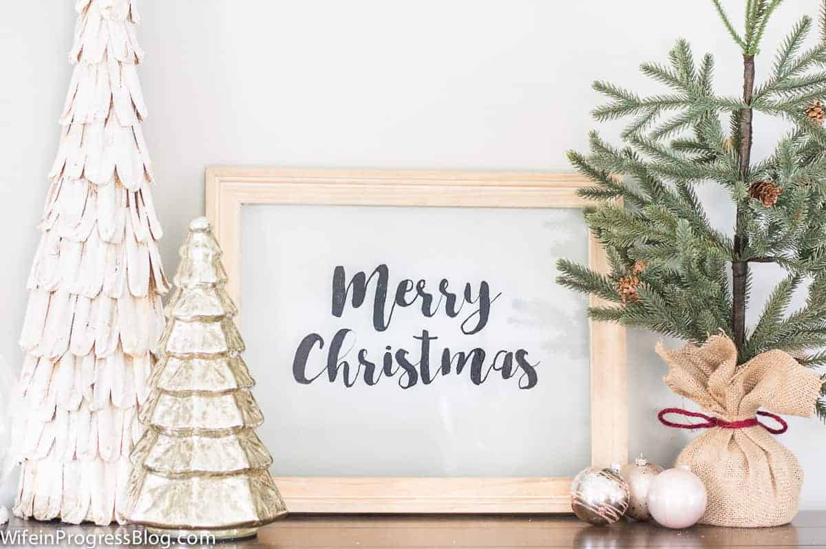 A \"Merry Christmas\" sign on clear glass, framed in unpainted wood, surrounded by ceramic Christmas trees and a green tree wrapped in burlap