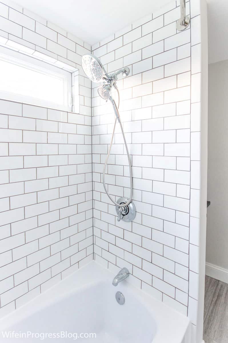 Chrome shower head, knob and faucet in bathtub area, surrounded by white subway tiles and gray grout 