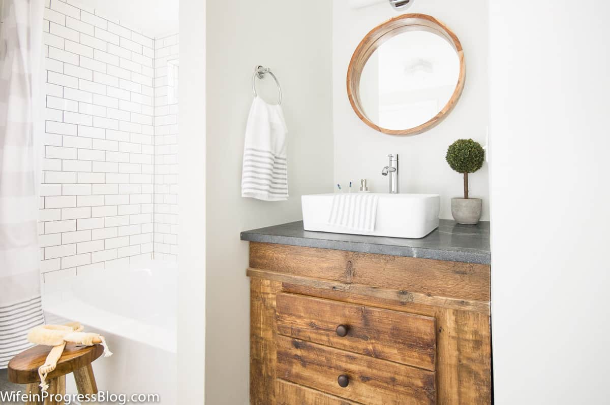 A rustic wooden vanity with a grey countertop and white, vessel sink with chrome faucet, and a framed, round mirror above