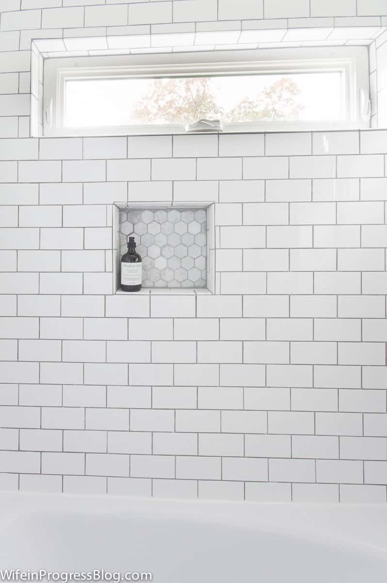 Medium gray grout white subway tile. Hexagon marble tile in shampoo niche.
Oblong horizontal window in top frame.