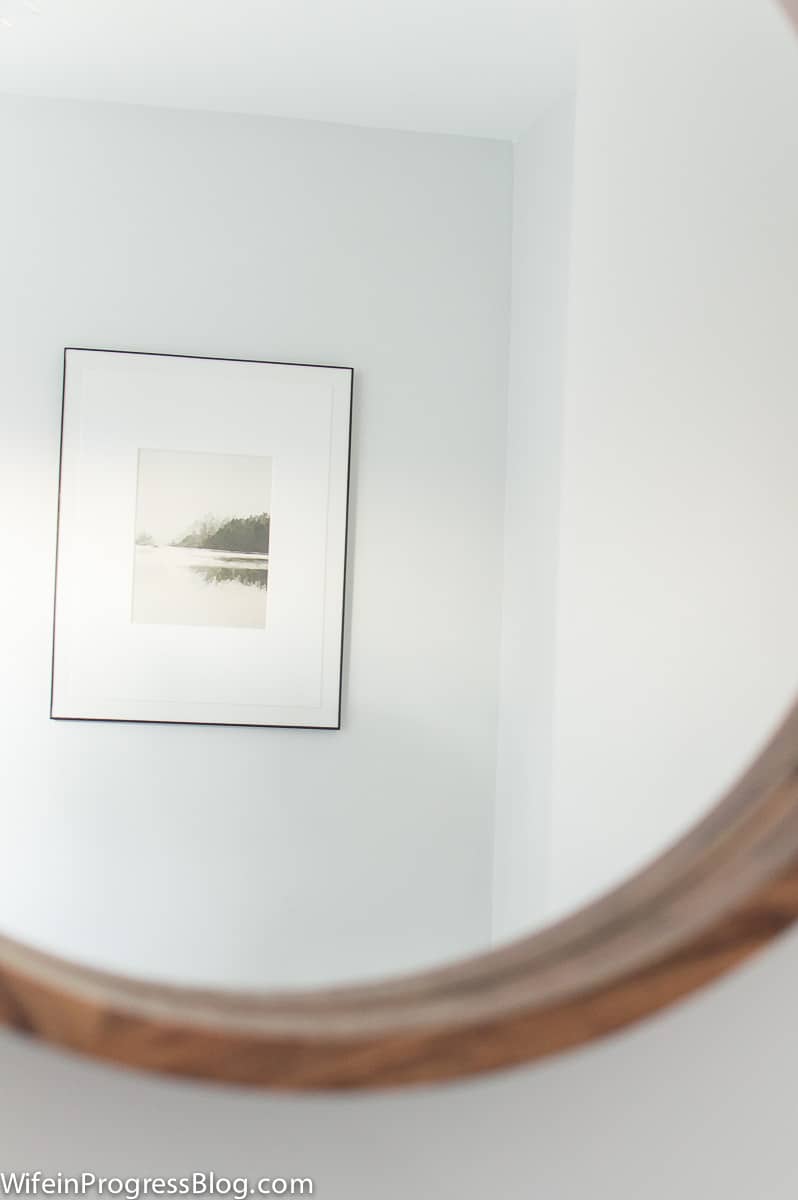 Reflection of framed landscape print through the round wood mirror are well-suited to a modern farmhouse bathroom