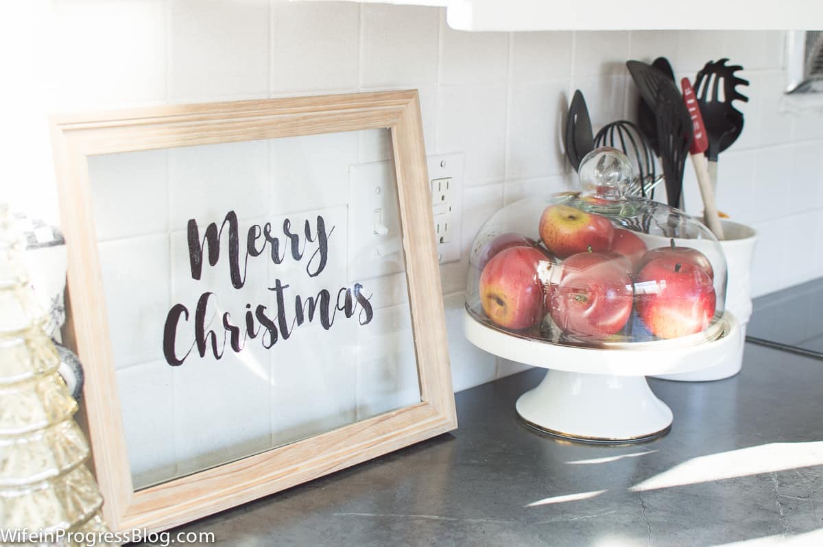 A \"Merry Christmas\" sign written on clear glass, framed in wood, next to a pedestal tray of apples covered by a glass dome