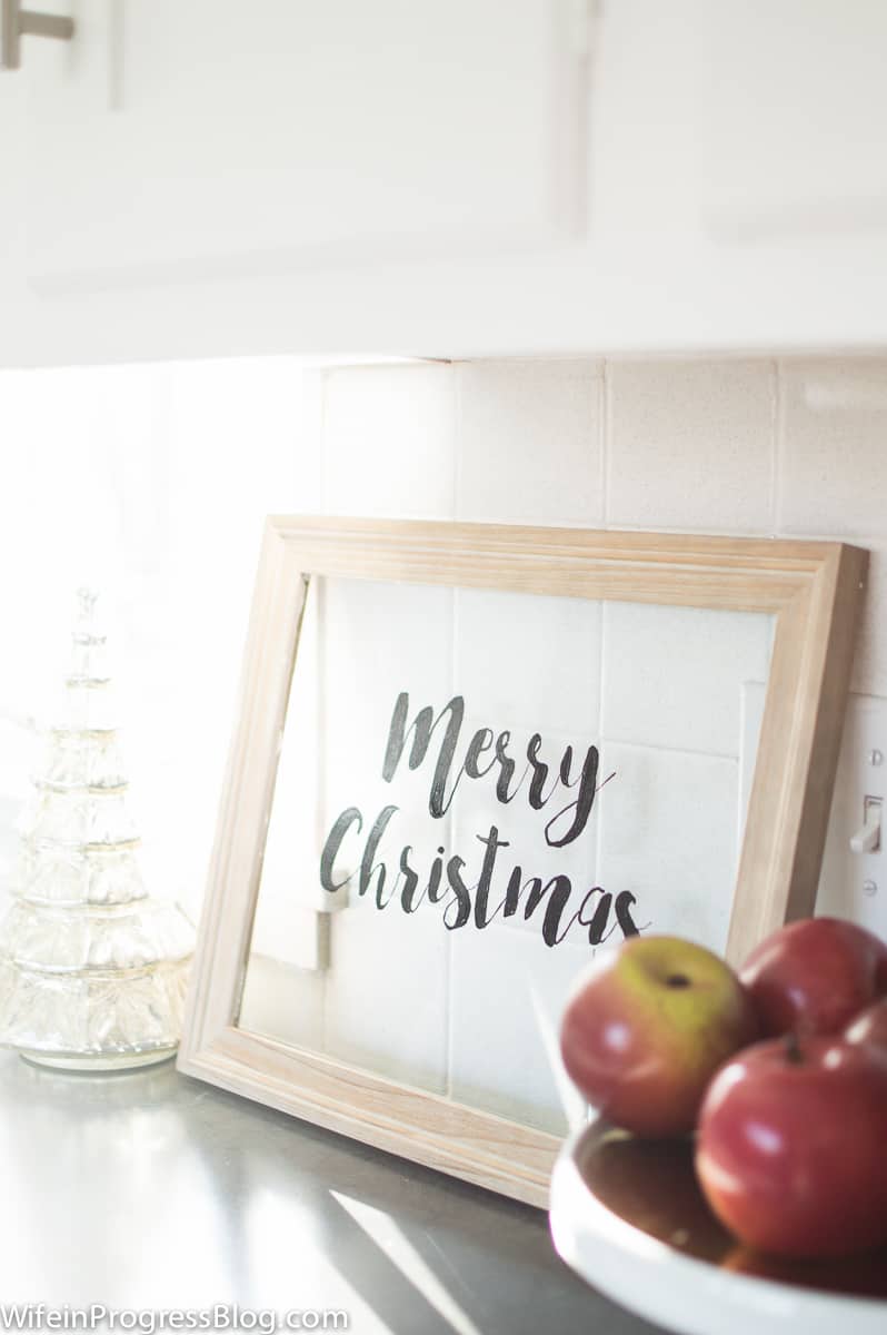 A small, white ceramic Christmas tree next to a \"Merry Christmas\" sign and a tray of apples