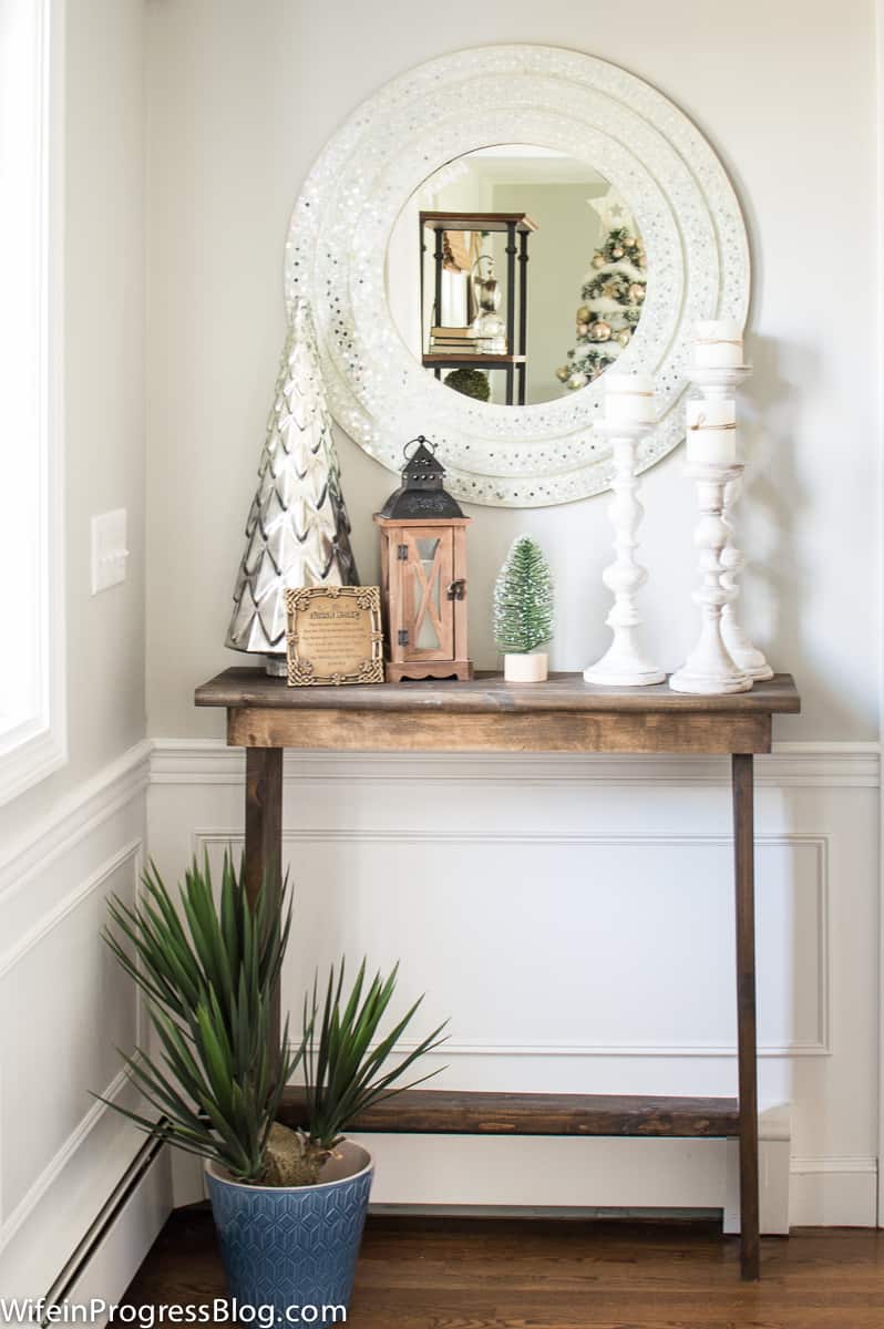 A console table holding Christmas decor, with a spiky potted plant below in a blue pot, and a round mirror above with a silver frame