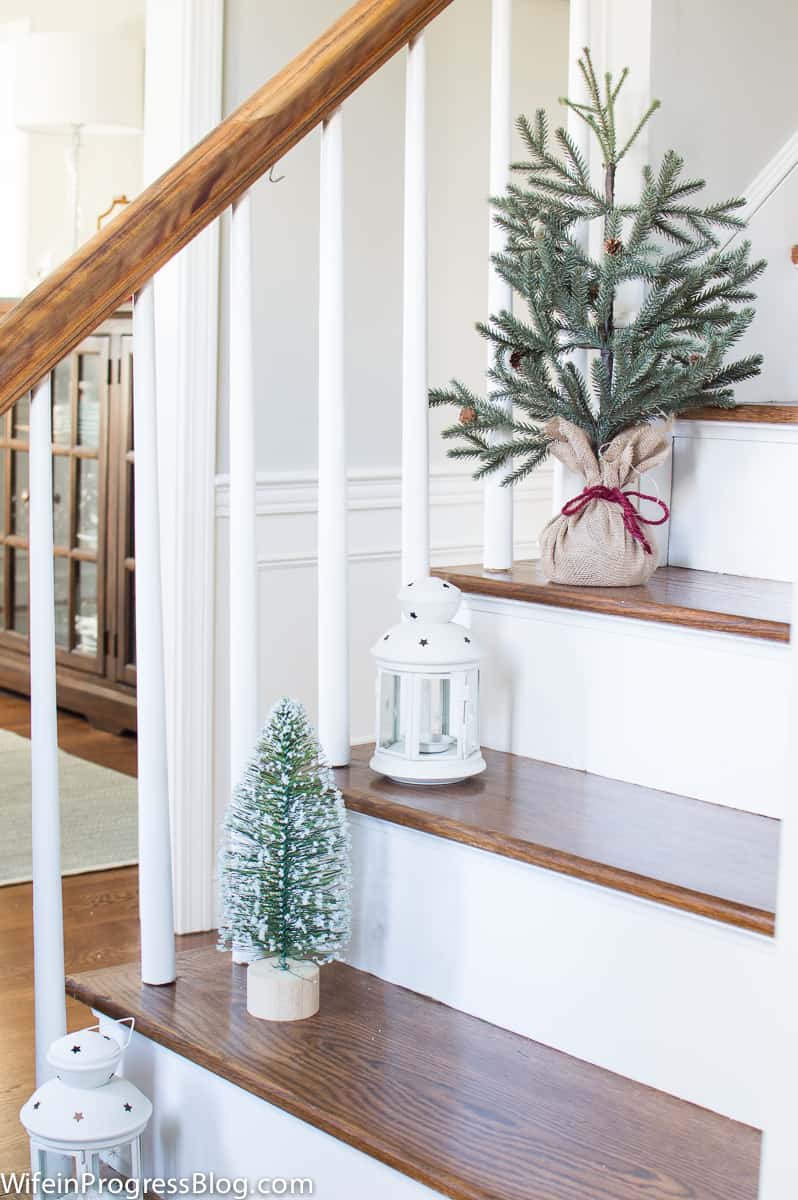 A wooden staircase with holiday items placed on the left side: a small tree wrapped in burlap and red twine, two white lanterns and a smaller tree