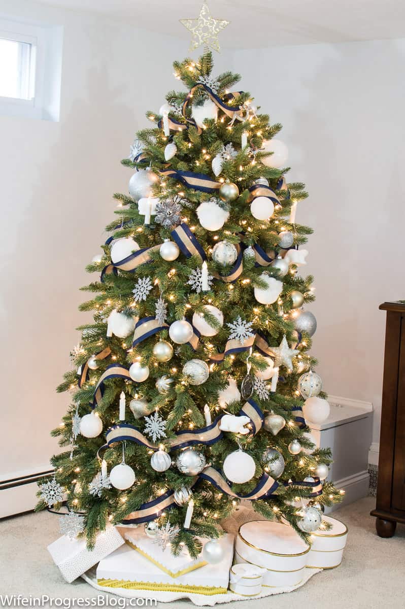 A lit Christmas tree, with white and silver ornaments and a blue and tan ribbon