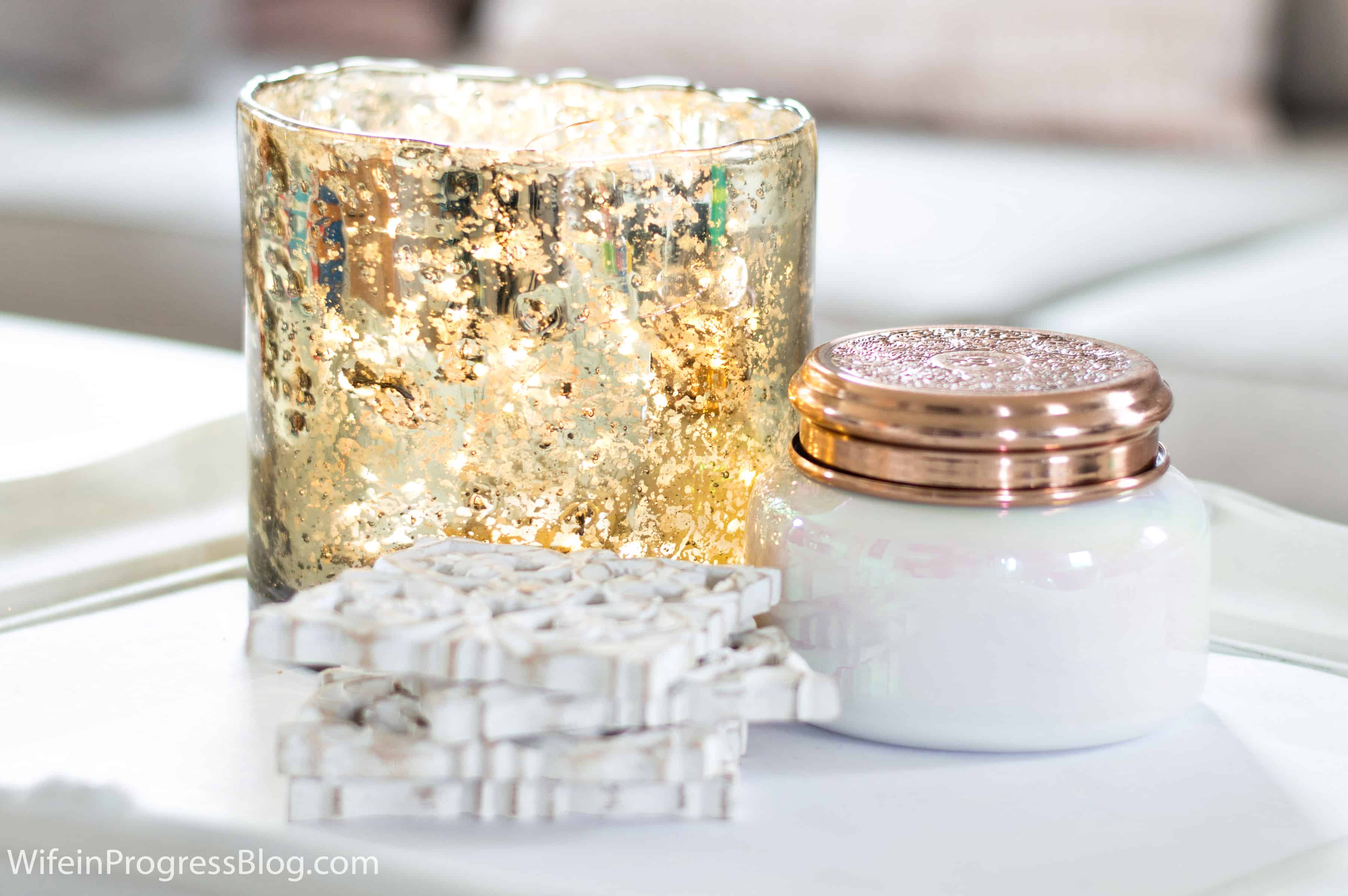 A gold leaf patterned candle holder, a white jar with copper cover, and a set of square white coasters with intricate pattern