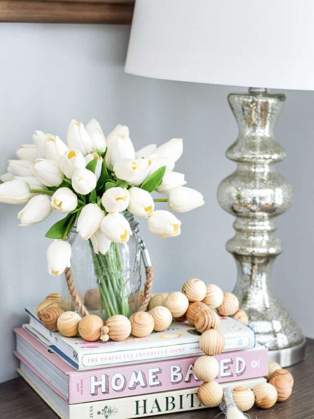 Tulips in a rustic glass vase on top of a stack of books, with a garland of small, wooden balls, next to a silver lamp