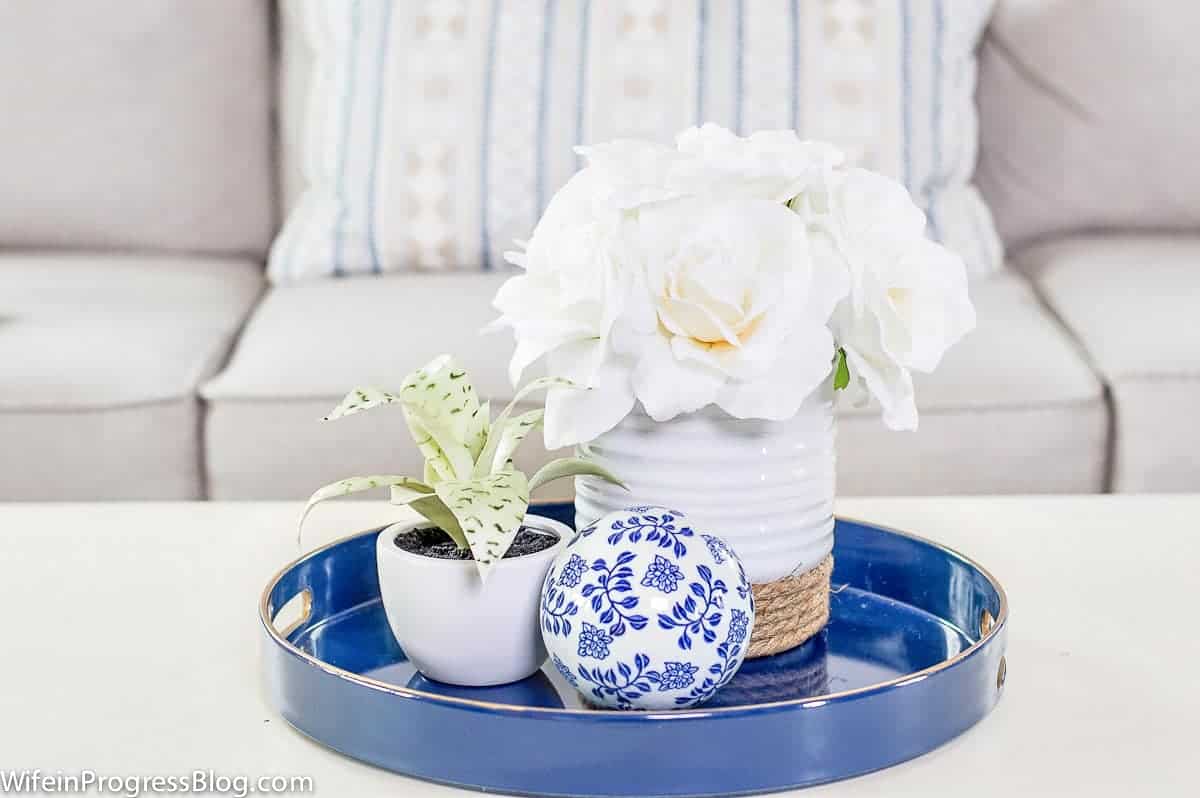 A short, white vase with large, white flowers, a blue and white ceramic ball and a small plant in a white pot, all resting on a round, blue tray.