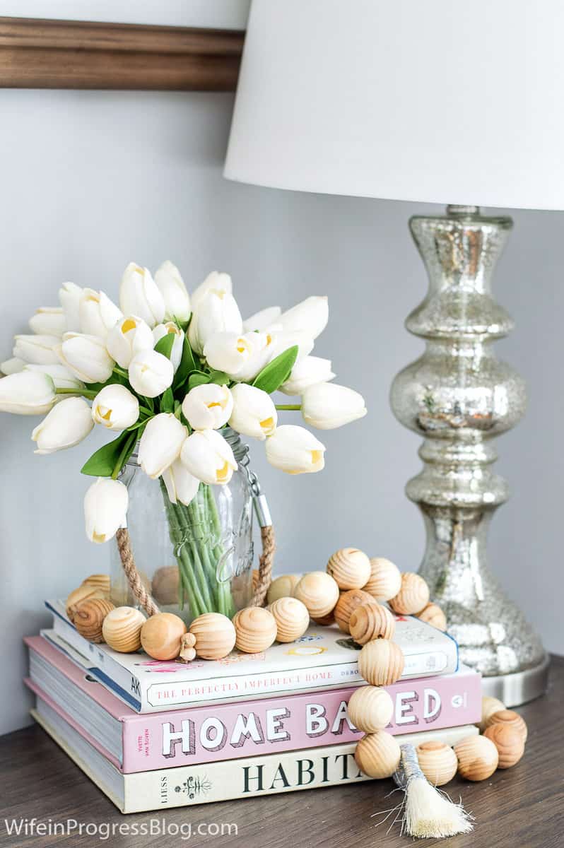 Tulips in a rustic glass vase on top of a stack of books, with a garland of small, wooden balls, next to a silver lamp