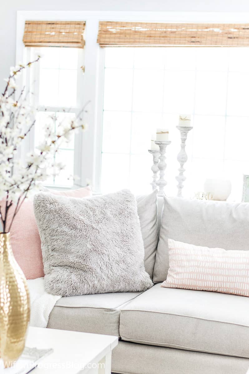 A large, plush grey pillow among light pink pillows, on a sofa, near a table with a slender gold vase holding tall white flowers