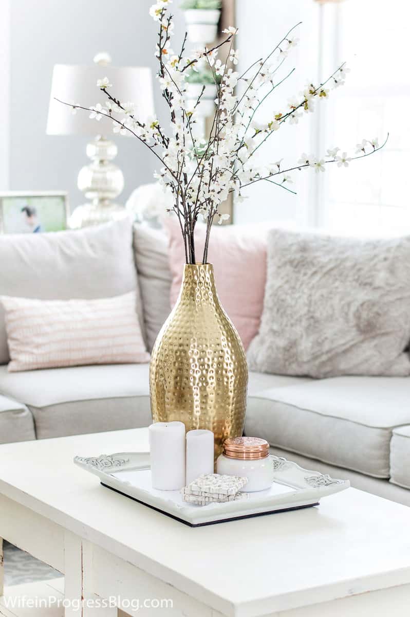A textured golden vase of flowers, resting on a square, white tray on an off-white coffee table in front of a light grey sofa