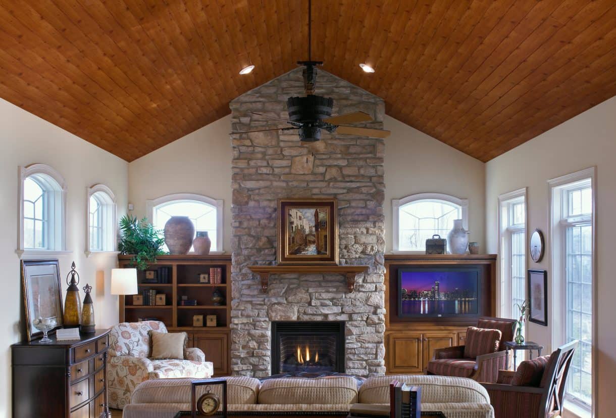 Natural wood stained planks in vaulted ceiling add character to tall stone fireplace wall and paned windows