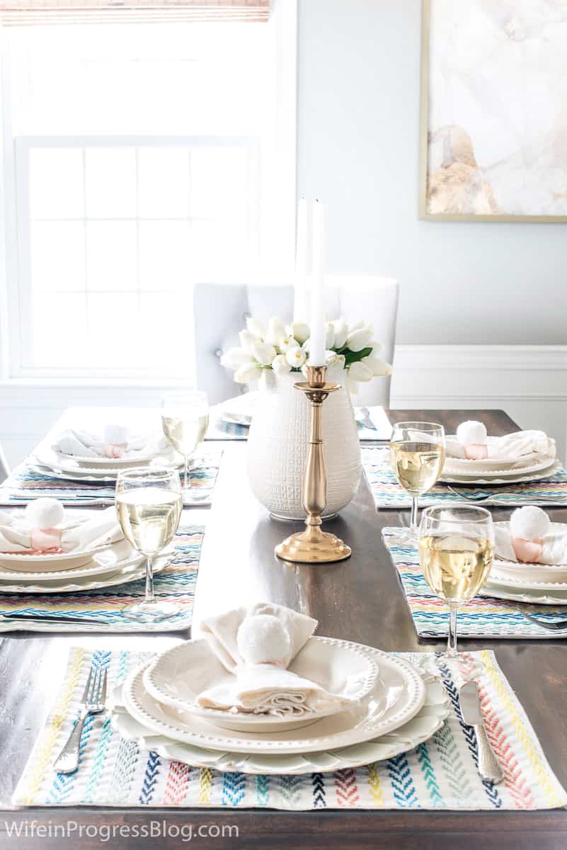 A tablescape with off-white plates and napkins, clear glass stemware, gold candle holders and a white vase with flowers