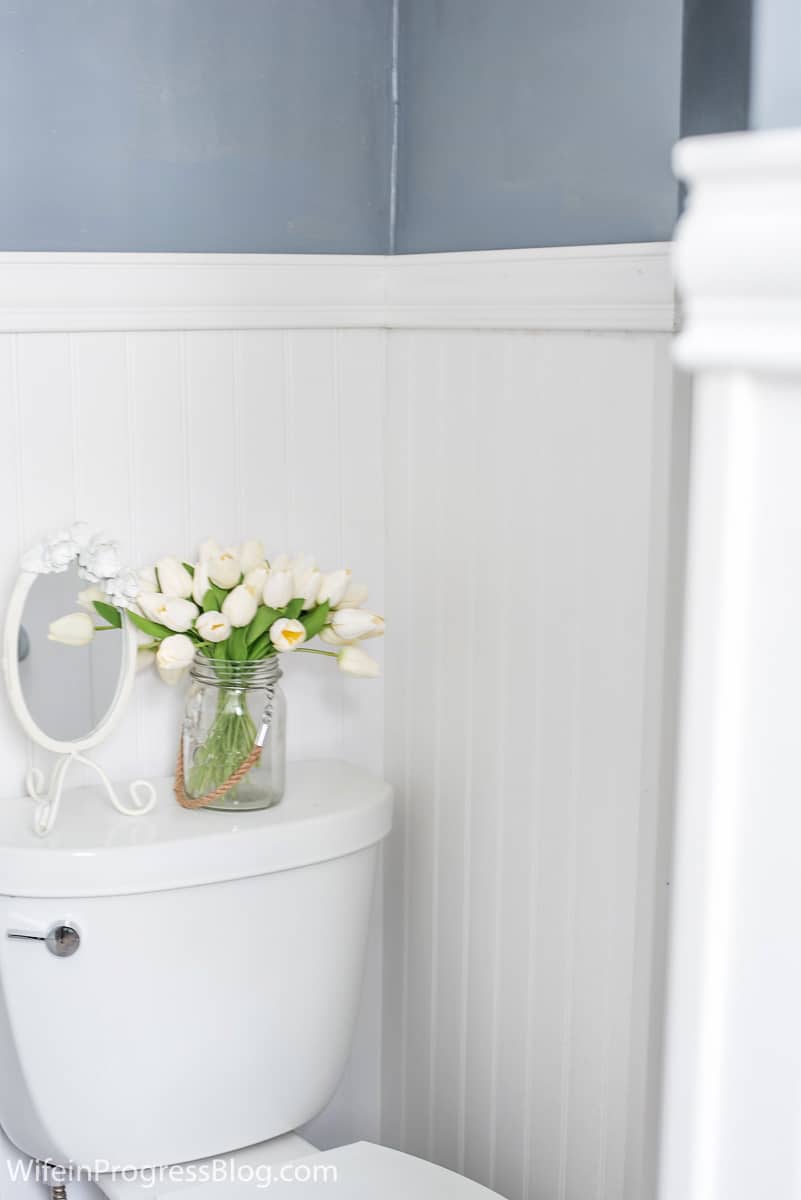 The finishing touches - antique mirror and vase of white flowers sit on toilet, framed by new beadboard wainscoting