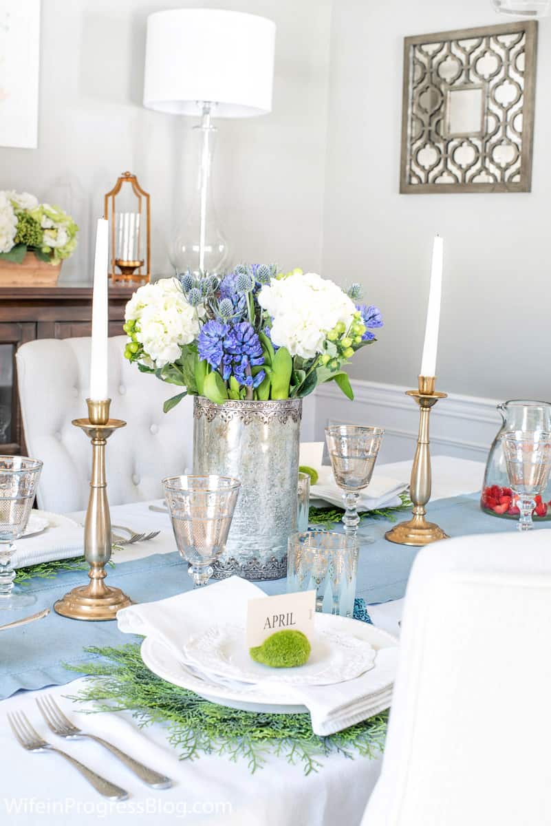 A tablescape with a blue runner, gold candle holders, a silver metal vase holding blue and white flower and natural green chargers 