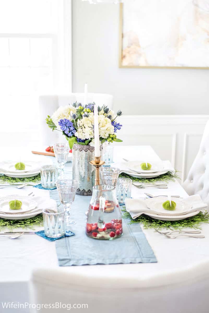 A tablescape with blue and white floral centerpiece, a light blue table runner and a glass pitcher of strawberry-infused water