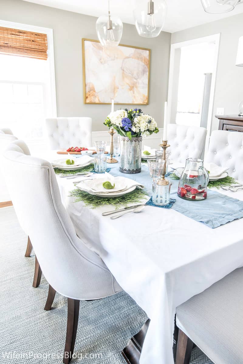 A tablescape with a light blue runner on top of a white tablecloth, and 6 white upholstered high-back chairs