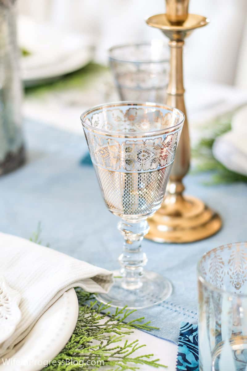 A wine glass with gold detailing on a light blue table runner, with a gold candlestick in the background