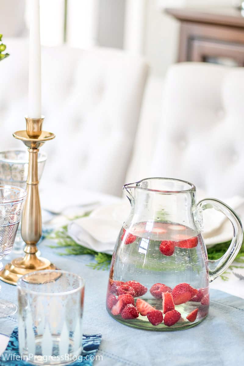 A clear glass pitcher of water with strawberries, near a golden candlestick and glass tumbler with gold detailing