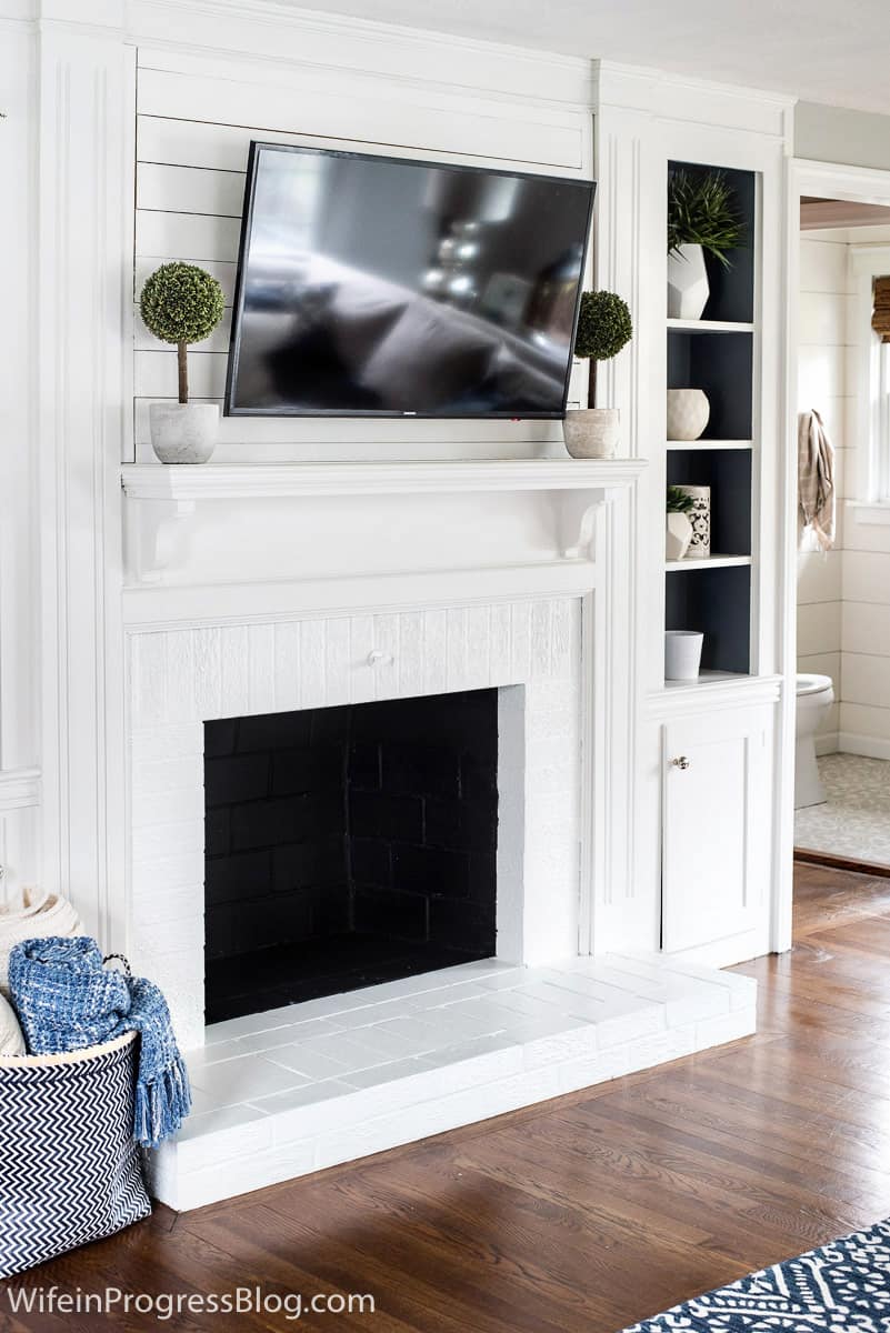 White fireplace, with white mantel and television overhead, topiary plants on either side of mantel