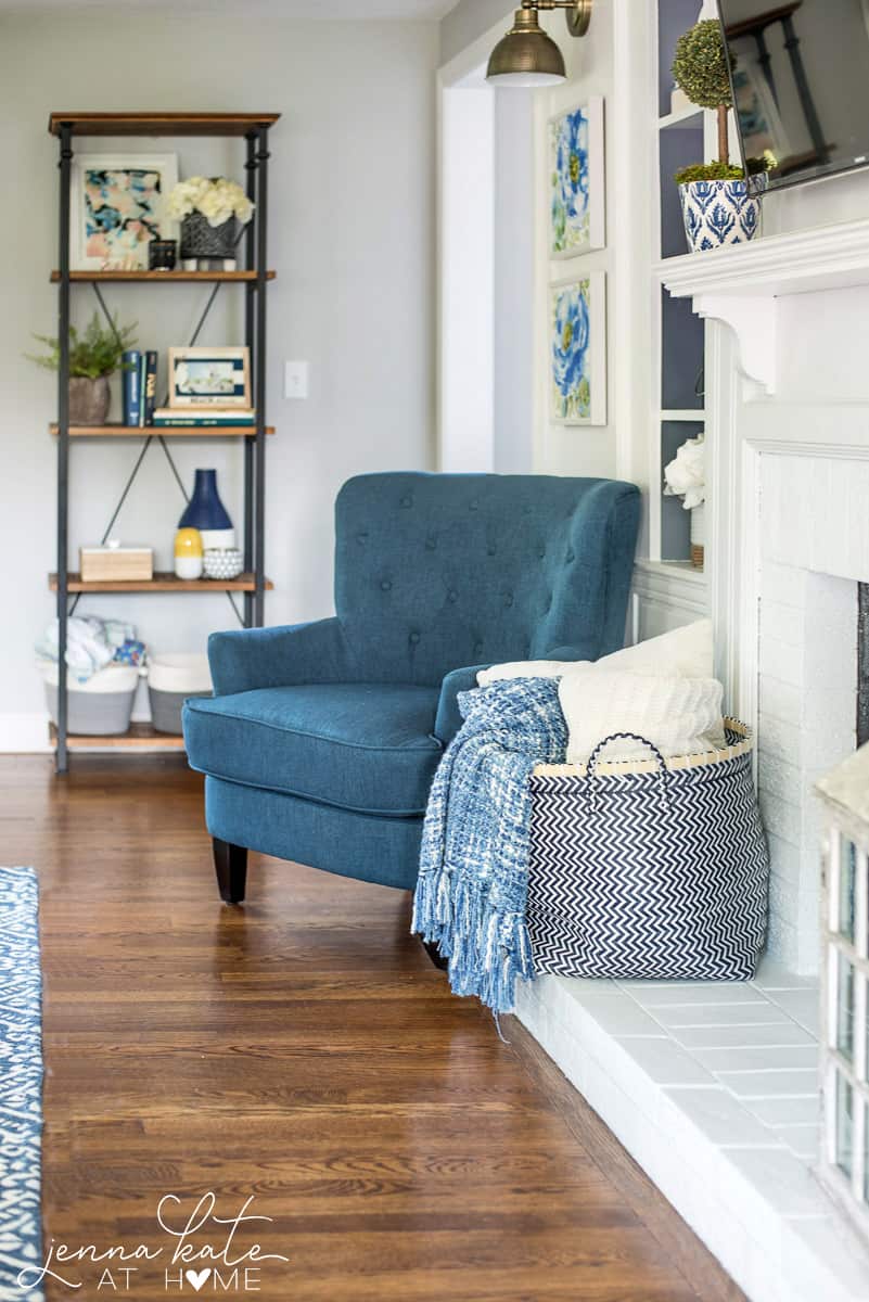 A living room with a tall, blue chair, a container of pillows and throws and a 4-tier shelf in the background with various decor items