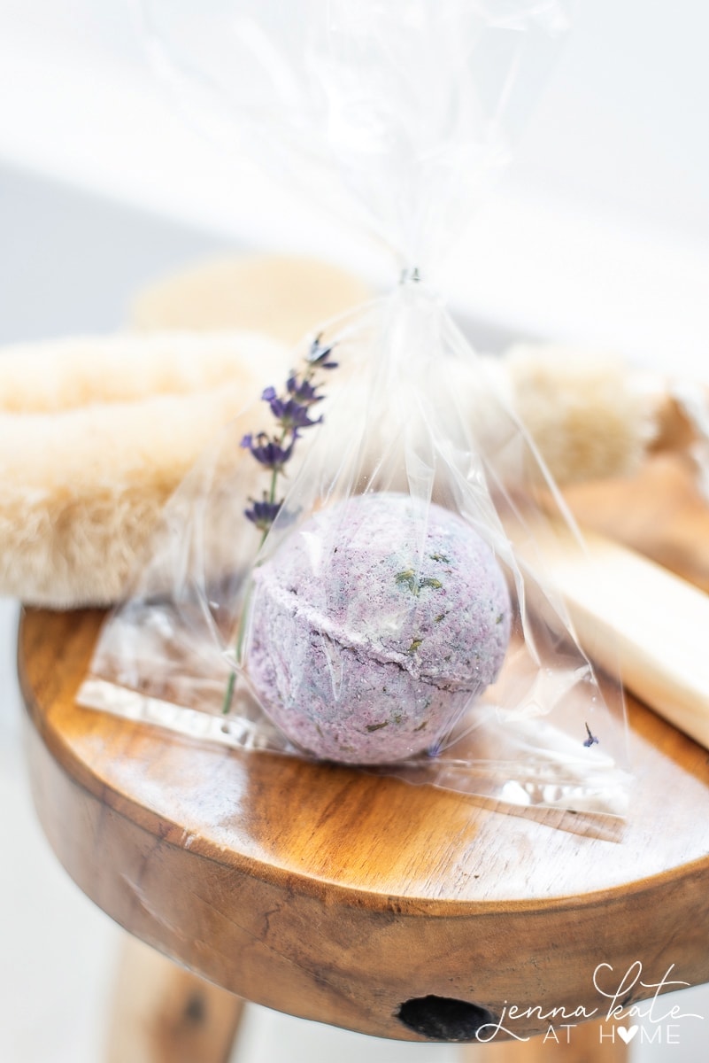 Purple bath bomb wrapped in cellophane bag with sprig of lavender, next to bath accessories