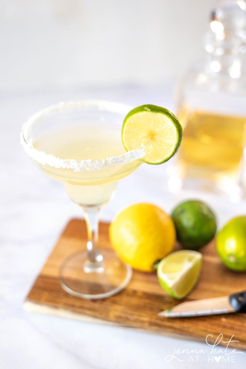 A drink in a margarita glass, resting on a wooden board with lemons and limes nearby